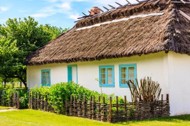 Mavi pencereli ve sazdan çatılı beyaz bir ev. Ev ahşap bir çitle çevrili ve önünde bir bahçe var.