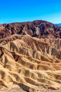Arka planda mavi gökyüzü olan bir dağ sırası. Dağlar kahverengi ve kayalıktır. Gökyüzü açık ve parlak