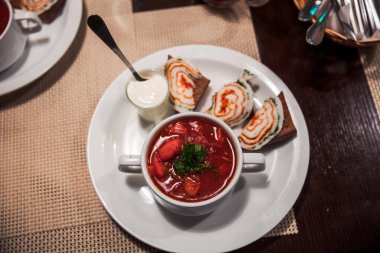 Bir kase çorba, bir kaşık ve beyaz tabakta küçük bir bardak sos. Tabak, üzerinde bez olan bir masanın üzerinde.