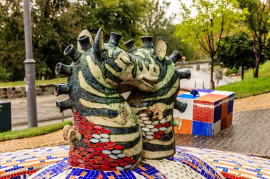 İki zebra heykeli bir çeşmenin üzerinde duruyor. Heykeller seramikten yapılmış ve parlak renklerle boyanmış. Çeşme bir park ile çevrili ve yakınında bir bank var. Sahne çok canlı.