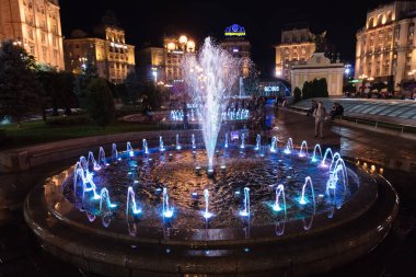 Geceleri mavi ışıkları olan büyük bir çeşme parlıyor. Çeşme bir park ve şehir sokağıyla çevrili.