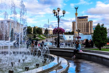 Bir kadın çeşmenin önündeki bankta oturuyor. Çeşme taştan bir geçitle çevrili ve bölgede yürüyen birkaç kişi var. Atmosfer sakin ve huzurlu.