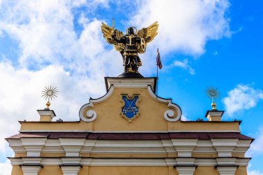 Bir binanın tepesine tünemiş bir melek heykeli. Heykel mavi ve altın bir dizaynla çevrilidir. Gökyüzü açık ve mavi, birkaç bulut etrafa saçılmış.