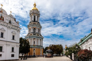 Altın kubbeli büyük, süslü bir bina beyaz bir binanın önünde duruyor. Gökyüzü mavi ve etrafta yürüyen birkaç kişi var.