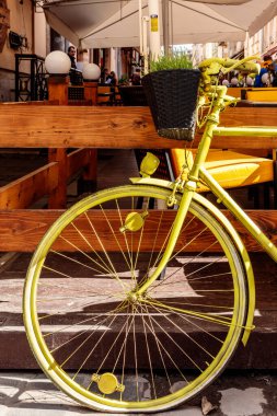 Önünde sepet olan sarı bir bisiklet ahşap korkuluklara yaslanıyor. Sepet saksı bitkisiyle dolu. Sahne bir restoranda ya da kafede geçiyor, sandalyeli.