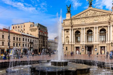 Meydanın ortasında bir çeşme. Etrafında insanlar dolaşıyor. Çeşme tuğla bir geçitle çevrili ve etrafında yürüyen birkaç kişi var.
