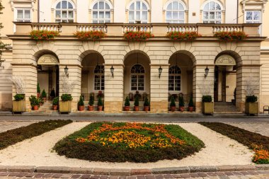 Önünde avlu olan büyük bir bina. Avlu çiçekler ve bitkilerle dolu ve kaldırımda birkaç saksı bitkisi var. Binanın bir sürü penceresi var.