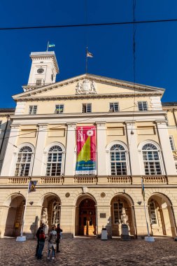Önünde pankart olan büyük bir bina. Pankart kırmızı ve beyaz. Binanın dışında bekleyen insanlar var.