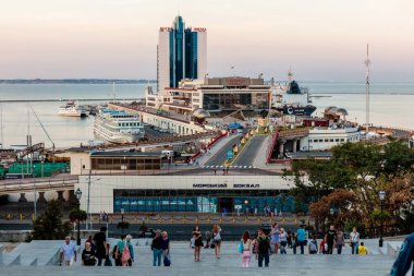 Bir grup insan büyük bir binanın önündeki merdivenleri çıkıyorlar. Sahne, el çantaları ve sırt çantaları taşıyan birçok insanın olduğu hareketli ve hareketli. Atmosfer çok enerjik.
