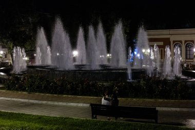 A fountain with many water spouts is lit up at night. A couple is sitting on a bench near the fountain clipart