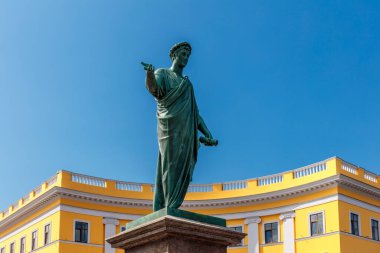 Sarı bir binanın önünde duran bir adam heykeli. Heykel bronzdan yapılmıştır ve uzun bir bornoz giymektedir. Bina yüksek ve bir sürü penceresi var. Gökyüzü açık ve mavi