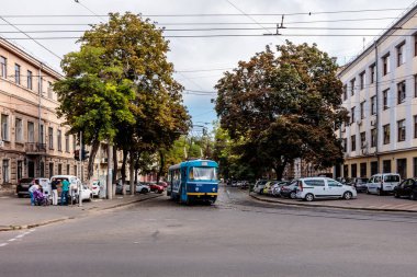 Mavi ve sarı bir tren bir şehirde bir caddede ilerliyor. Tren arabalar ve insanlarla çevrili ve kaldırım boyunca birkaç bank var. Sahne canlı ve hareketli.