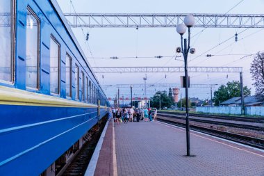 Platformda duran insanların olduğu mavi bir tren. Tren bir istasyonda ve insanlar onu bekliyor.