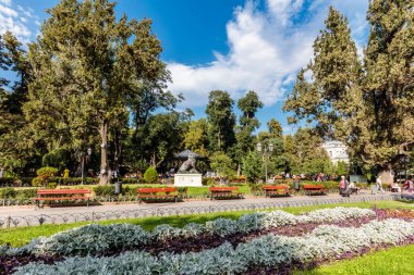 Heykeli ve bankları olan bir park. Heykel bir adamın ve banklar kırmızı. Parkta bir sürü insan var, bazıları banklarda oturuyor ve diğerleri etrafta dolaşıyor.