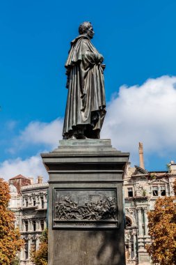 Bir binanın önünde duran bir adam heykeli. Heykel bronzdan yapılmıştır ve çok ayrıntılıdır. Gökyüzü mavi ve arka planda bazı bulutlar var