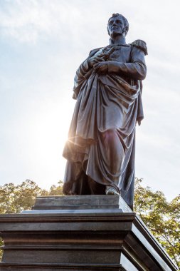 Omuzlarının üzerinde pelerin olan bir heykelin üzerinde duran bir adam heykeli. Heykel ağaçlarla çevrili ve gökyüzü mavi.
