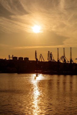 Güneş suyun üzerinde batıyor, arka plandaki binalara sıcak bir ışıltı saçıyor. Gökyüzünün renklerini yansıtan suyla sahne huzurlu ve huzurlu.