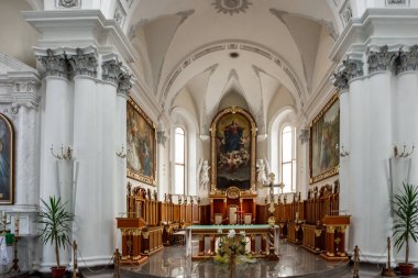 Bir kilisenin içi çok büyük ve çok beyazdır. Bir sürü saksı bitkisi ve birkaç heykel var. Saksıdaki bitkiler ön planda ve heykeller arka planda.