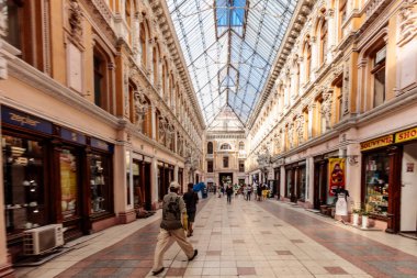 A long, narrow shopping mall with a blue roof. People are walking around and shopping. The mall is filled with many stores and a few potted plants clipart