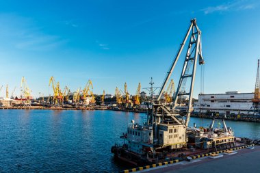 Suyun üzerinde büyük bir su kütlesinin yanında büyük bir vinç var. Vinç birçok gemi ve tekne ile çevrilidir. Sahne sakin ve huzurlu, su ve gökyüzü huzurlu bir zemin sağlıyor.