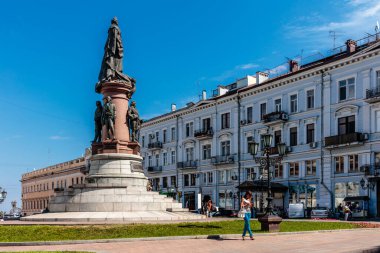Bir kadın büyük bir heykelin önünde yürür. Heykel bir park ve binalarla çevrili. Gökyüzü açık ve güneş parlıyor.
