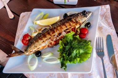 Balık, yanında sebze olan beyaz bir tabakta servis edilir. Balık ızgara ve üstünde limon dilimi var. Sebzeler arasında marul, domates ve soğan var.