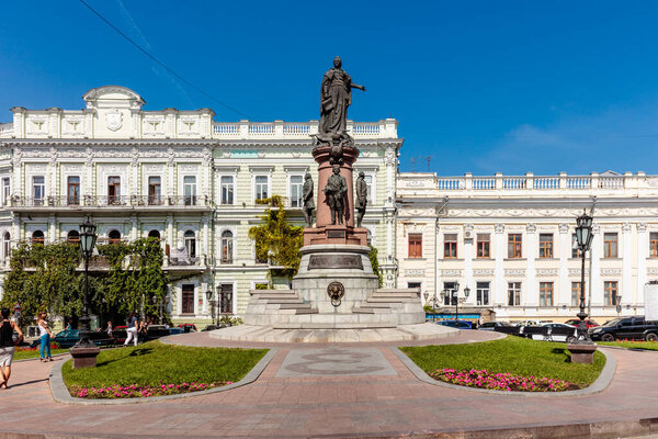 Статуя мужчины стоит перед зданием. Статуя окружена парком и тротуаром. Небо чистое и голубое.