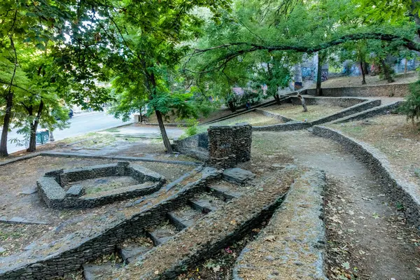 Ortasında taş bir çeşme olan taş bir patika. Geçit ağaçlarla çevrili ve yan tarafında taş bir duvar var.