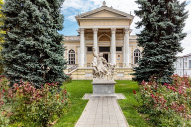 Önünde insan ve hayvan heykelleri olan büyük bir bina. Heykeller, çocuklu bir adam ve bir kadının ve bir atın heykelleri. Bina ağaçlarla çevrili ve önünde bir bahçe var.