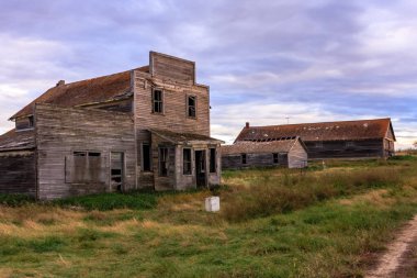 Küçük, yıkık dökük bir ev arka planda birkaç evle birlikte bir tarlada duruyor. Gökyüzü bulutlu ve atmosfer sakin ve huzurlu.
