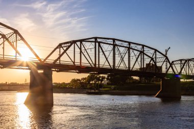 Bir köprü, üzerinde güneş parlayan bir nehir boyunca uzanır. Köprü eski ve kırsal bir görünüme sahip.