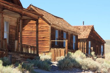 Bir sürü penceresi ve verandası olan üç eski ahşap ev. Evler bir tepenin üzerinde ve etraflarında çimenler büyüyor.