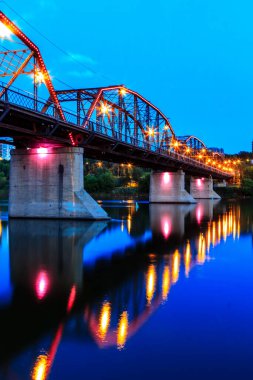 Üzerinde ışık olan bir köprü suya yansıyor. Köprüdeki ışıklar pembe ve mor.