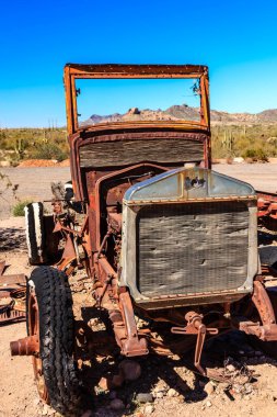 Eski, paslı bir kamyon çölde park halinde. Kamyon paslanmış ve soluk turuncu bir boyası var. Çöl manzarası çorak ve ıssız, yaşam belirtisi yok.