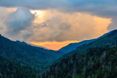 Gökyüzü bulutlarla dolu ve güneş batıyor. Arkadaki dağlar ağaçlarla kaplı.