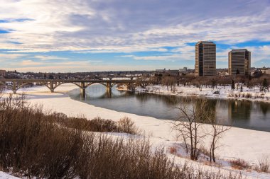 Nehir ve köprüsü olan karlı bir manzara. Gökyüzü bulutlu ve güneş batıyor.
