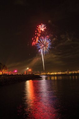 Gece gökyüzünde bir su kütlesinin üzerinde havai fişek gösterisi yakılıyor. Havai fişekler kırmızı, beyaz ve mavi ve havaya fırlıyorlar. Huzurlu ve huzurlu bir sahne.