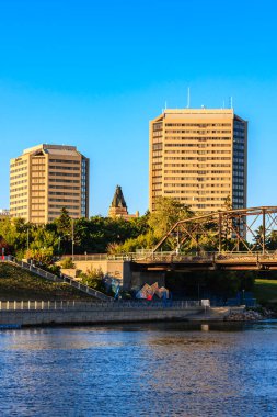 Bir köprü iki yüksek binanın önündeki bir nehri kapsar. Gökyüzü mavi ve güneş batıyor.