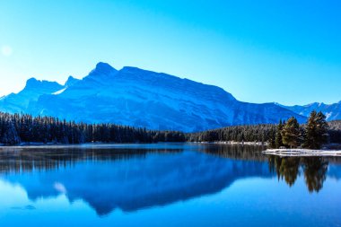 Bir dağ sırası bir gölün suyuna yansıtılır. Gökyüzü açık ve mavi ve dağlar karla kaplı. Sahne huzurlu ve sakin, dağlarla birlikte.