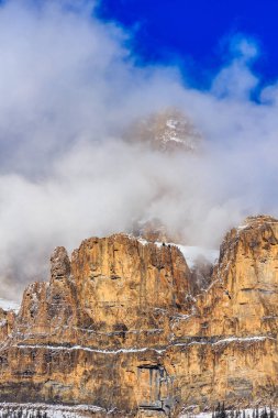 Kar ve bulutlarla kaplı bir dağ sırası. Gökyüzü mavi ve bulutlar beyaz. Dağlar yüksek ve kayalıktır.
