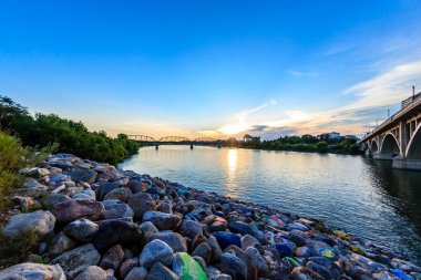Bir köprü arka planda güzel bir günbatımı olan bir nehri kapsar. Su sakin ve gökyüzü berrak.