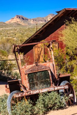 An old, rusted car is sitting in front of a red house. The car is surrounded by weeds and grass, giving it a somewhat abandoned and neglected appearance clipart