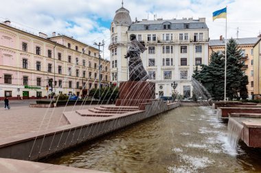Şehir meydanındaki bir çeşmenin önünde bir adam heykeli var. Çeşme bir taş duvar ve birkaç ağaçla çevrilidir. Huzurlu ve huzurlu bir sahne, etrafta insanlar geziniyor.