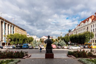 Bir binanın önünde duran bir adam heykeli. Heykel bir park ve işlek bir cadde ile çevrilidir. Gökyüzü bulutlu ve caddede park edilmiş bir sürü araba var.