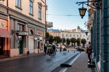 Kaldırımda dikilen bir adamın olduğu sokak sahnesi. Etrafta dolaşan birkaç kişi ve caddeye park etmiş birkaç araba var. Atmosfer canlı ve hareketli.