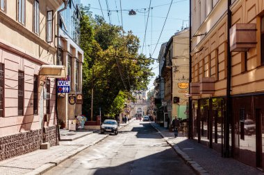 Arabası olan bir cadde ve yürüyen birkaç kişi. Sokak binalarla dolu ve bir sürü ağaç var.