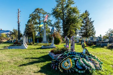 Yerde büyük bir çiçek aranjmanı olan bir mezarlık. Çiçekler çeşitli renklerdedir ve bir daire şeklinde düzenlenmişlerdir. Mezarlık ağaçlarla çevrili ve birkaç mezar taşı var.