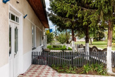 Üzerinde mavi ve sarı bir tabela olan beyaz bir ev. Evin etrafı çitlerle çevrili ve küçük bir bahçesi var.