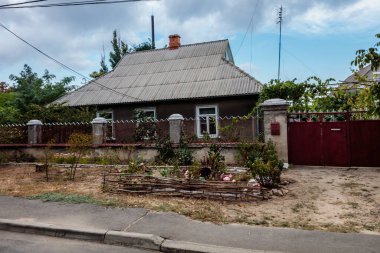 Kırmızı kapısı ve beyaz posta kutusu olan bir ev. Ev bir çitle çevrili ve küçük bir bahçesi var. Gökyüzü bulutlu ve cadde boş.