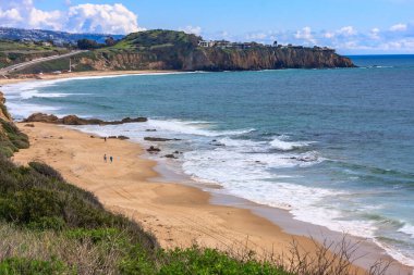 Kayalık bir kıyı şeridi ve mavi bir okyanusu olan bir sahil. Kumsal bomboş, kıyıda yürüyen iki kişi dışında.